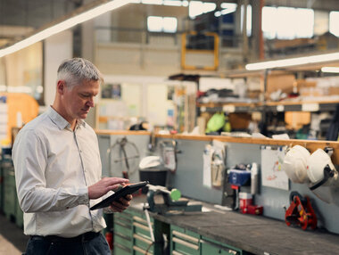 Für Mario Figura sind Ausbildung und Studium eine perfekte Kombination für eine Leitungstätigkeit im praktischen Bereich. Betreibsleiter steht mit Klemmbrett vor einer Werkbank